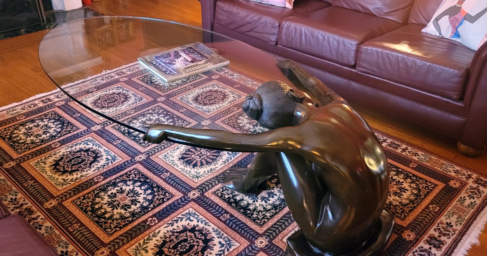 Bronze & Glass Coffee Table by Aug Moreau with a naked woman holding the glass knees raised to her chest, a on a multi colored rug in front of a burgundy leather couch