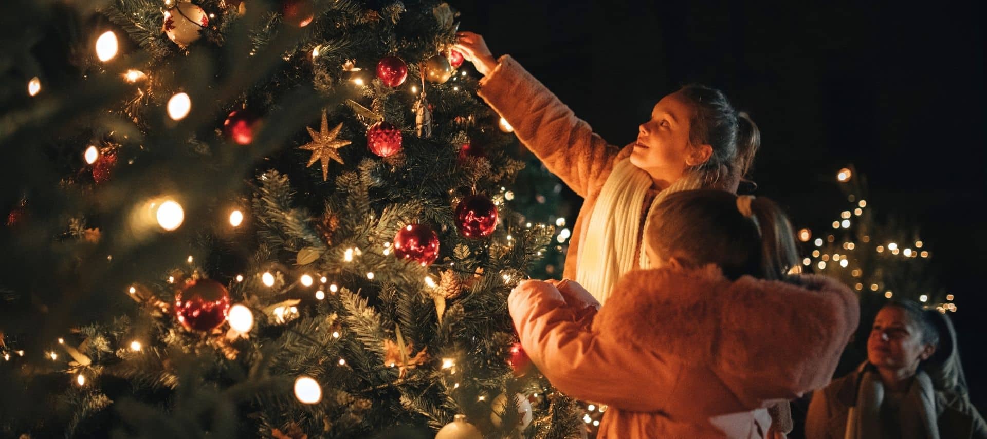 a smiling woman and two children admiring and decorating a christmas tree with lights and ornaments during the evening
