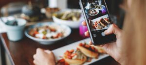 A person holds a phone taking a photo of plated food on a table, with dishes visible on the screen and in the background.