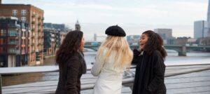 three friends laughing together while standing on a bridge overlooking the river and city skyline in winter