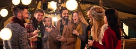 a group of friends gathered at a holiday event wearing scarves and coats laughing and holding drinks with warm lights glowing around them