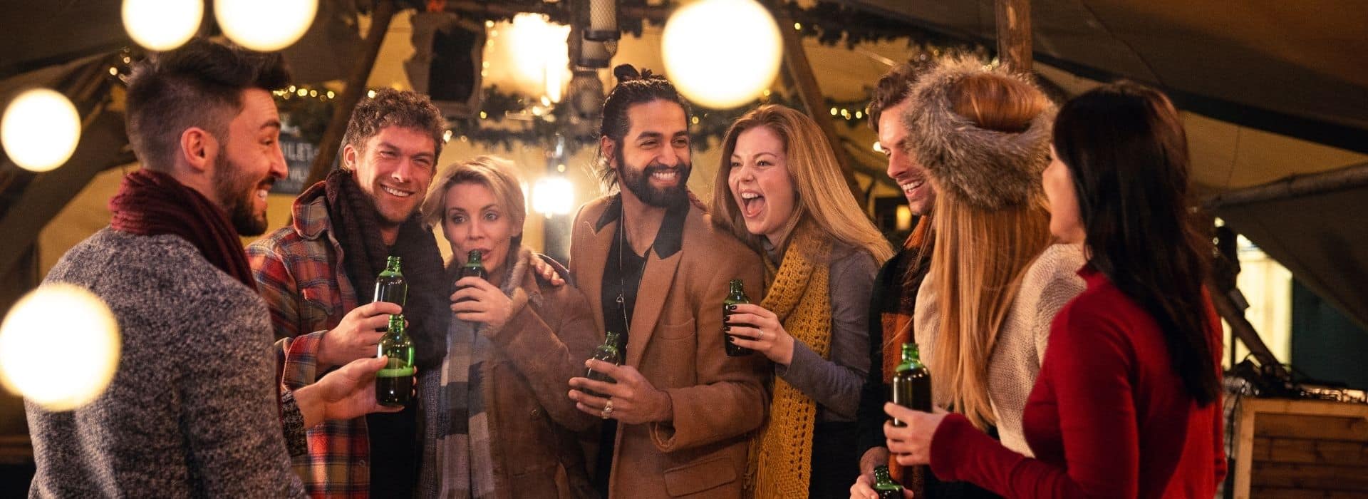 a group of friends gathered at a holiday event wearing scarves and coats laughing and holding drinks with warm lights glowing around them