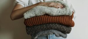person holding a folded stack of cozy knit sweaters in neutral and warm tones