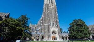 The towering Gothic architecture of Duke University stands against a bright blue sky, framed by large trees.
